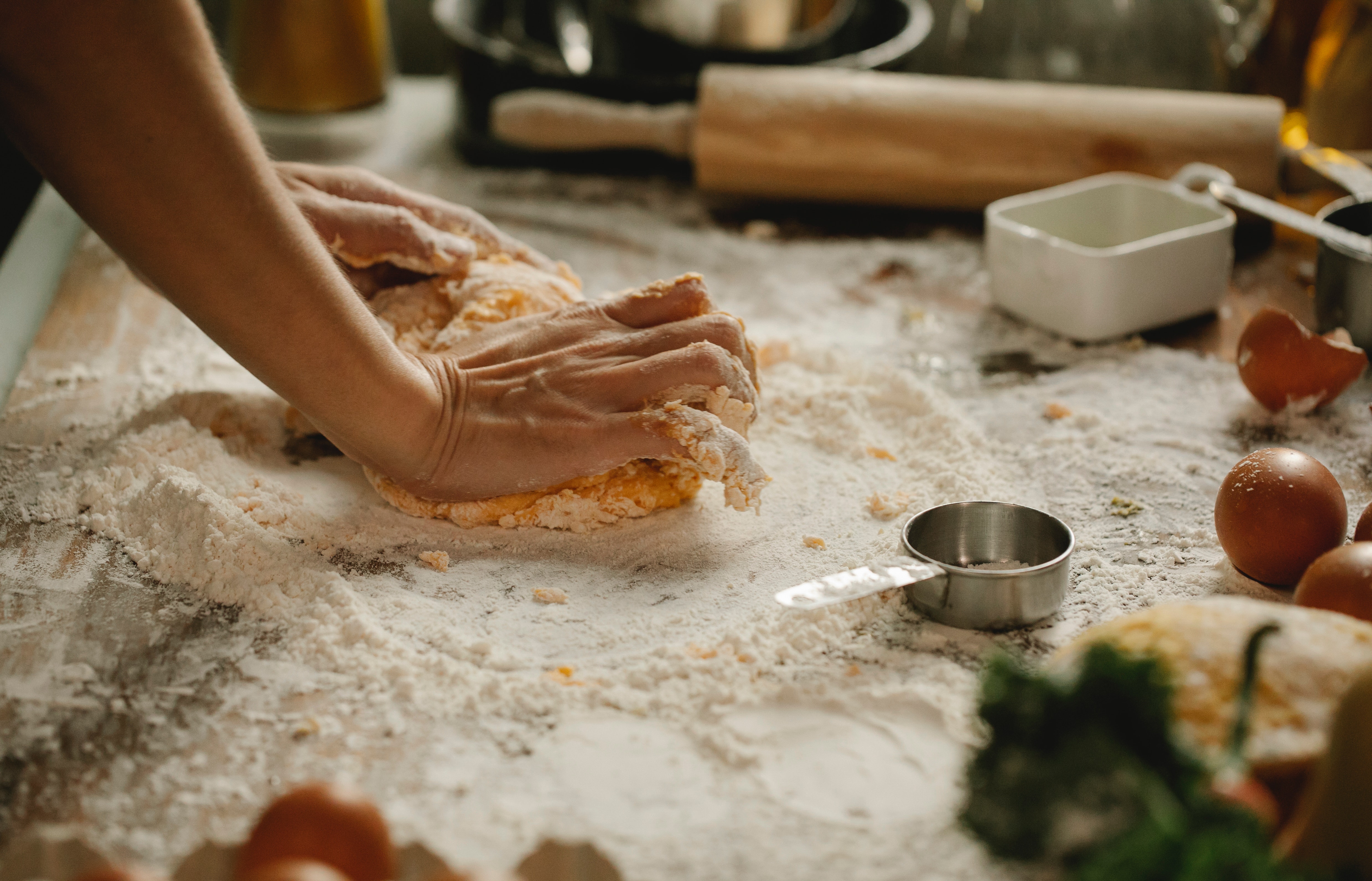 Hands working with dough on a floured board with a rolling pin, egg, and a measuring cup