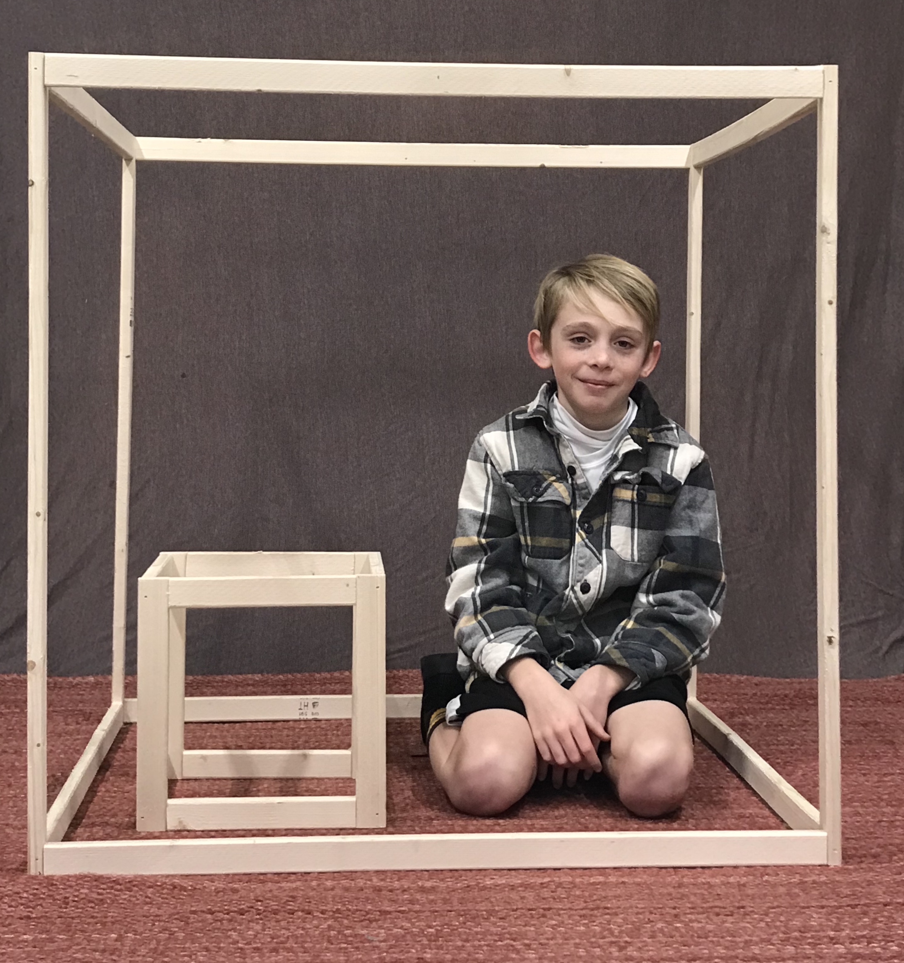 <p>A student and two rectangular prisms. . The student is sitting inside the large rectangular prism and is next to the smaller prism. </p>
