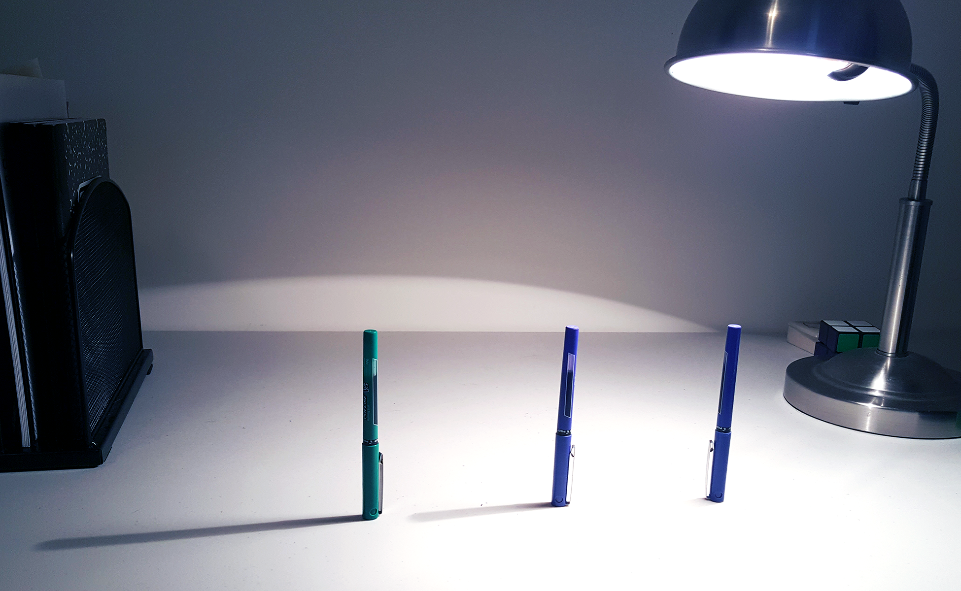 <p>Photograph of 3 pens standing upright. Lamp on right. Longest shadow formed by pen on far left, shorter shadow on middle pen, no shadow on pen on right.</p>
