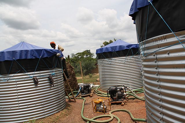 Photo of three water tanks with drainage hoses. A worker is on a ladder, leaned against the side of one of the tanks.
