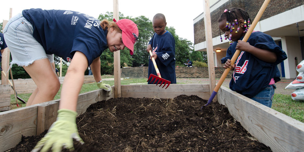<p>One adult and two children filling an outdoor square planter with soil</p>
