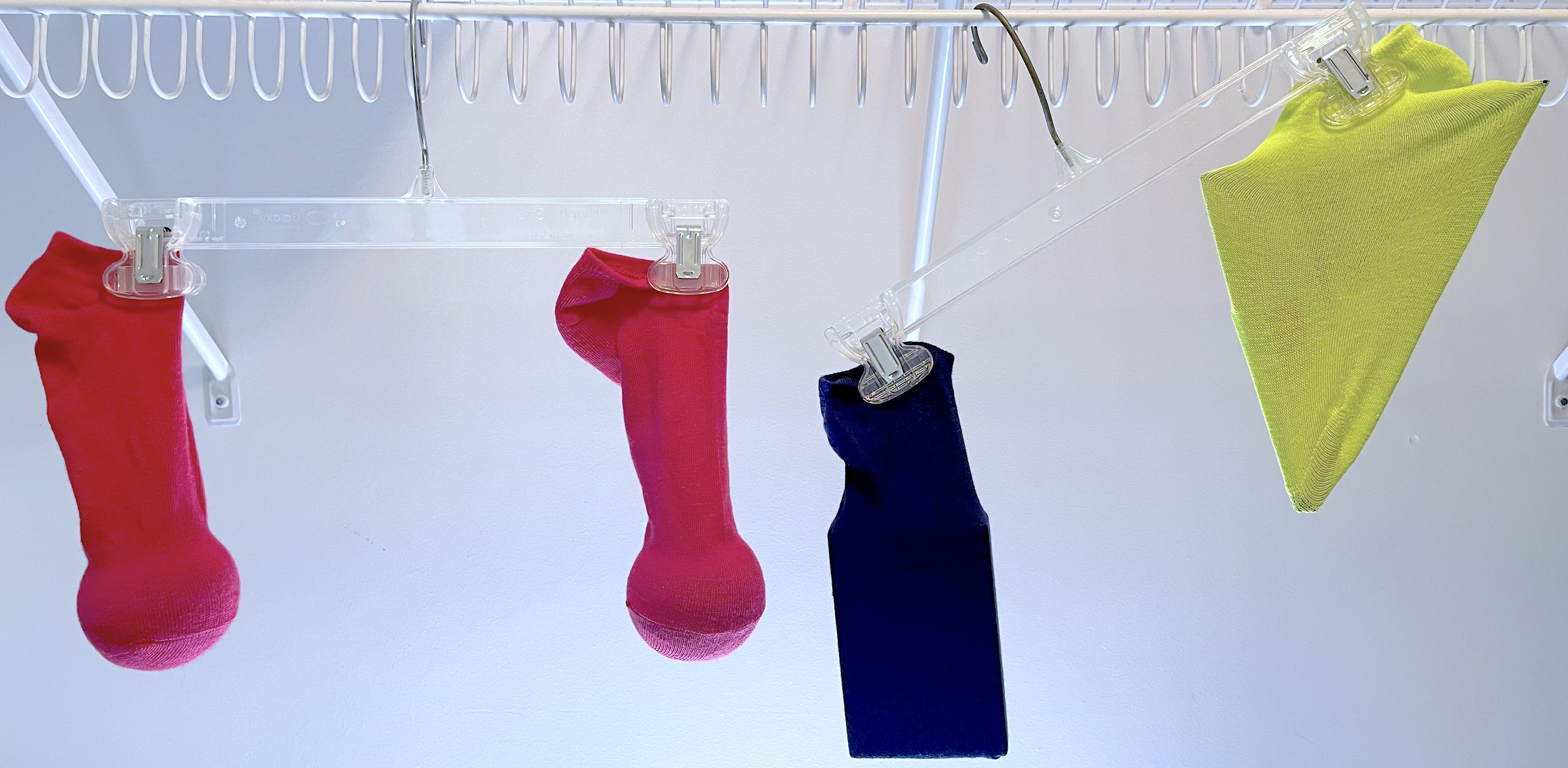 Socks with items inside hanging from hangers. The left hanger is balanced with 2 red socks that have balls inside. The right hanger is unbalanced. A navy sock has a rectangular object inside and the yellow sock has a triangle inside.